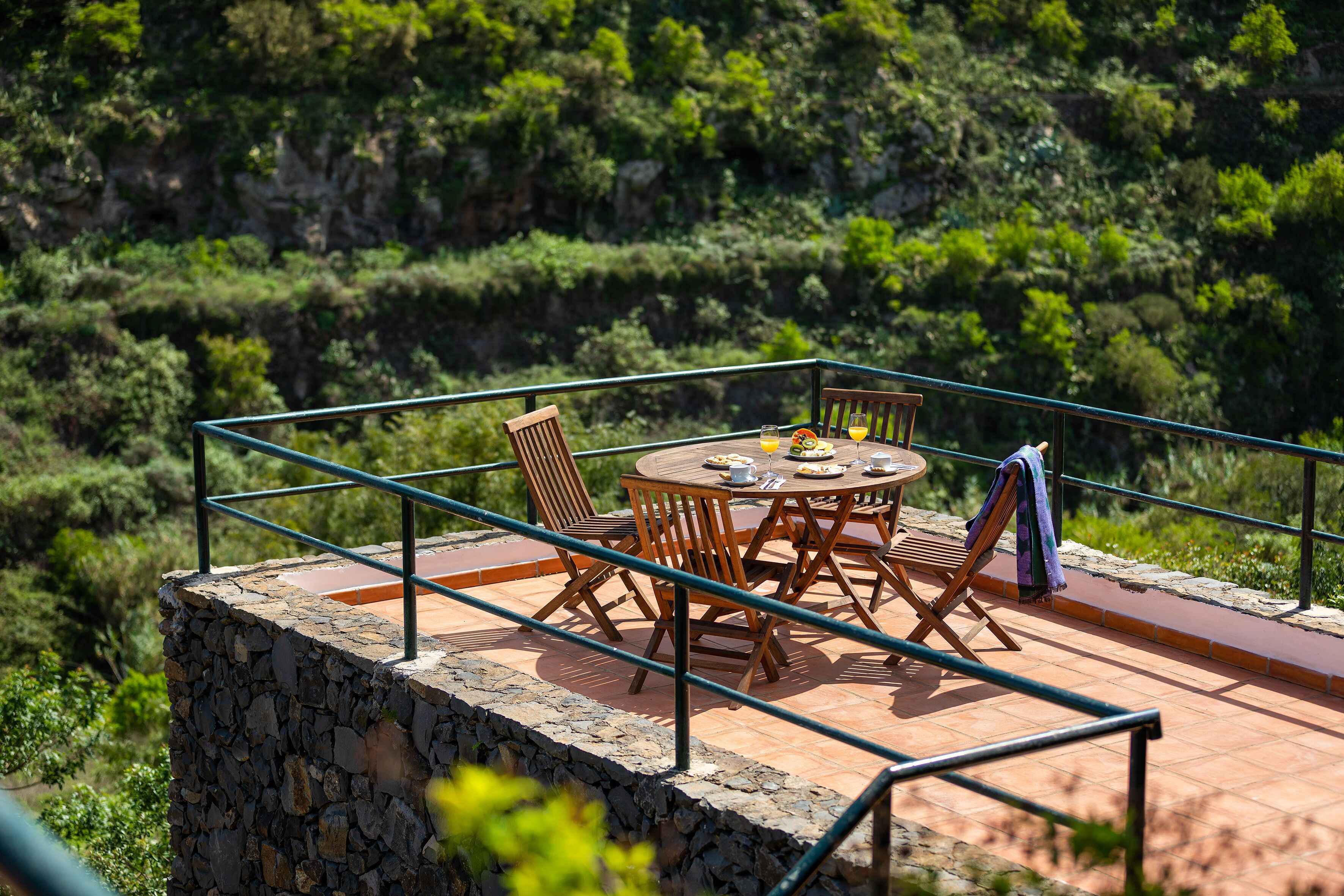 A terrace with a stone wall, wooden furniture, and a view of a lush green hillside.