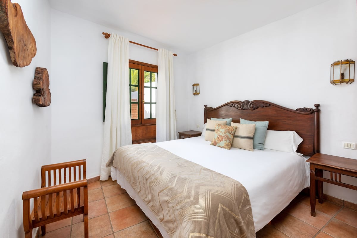 A bedroom with a wooden bed, a wooden nightstand, a chair, and a window with curtains.
