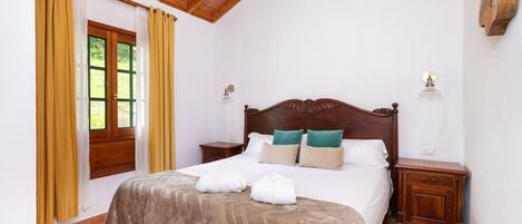 A bedroom with a wooden ceiling, a large bed with a patterned bedspread, two bedside tables, and a window with curtains.