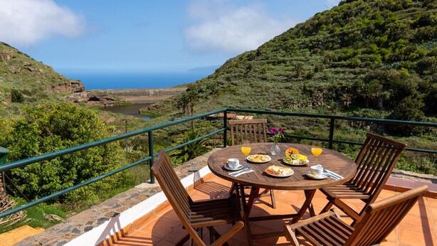 A terrace with a round wooden table and four chairs, overlooking a lush green landscape and the sea. - Las Casas del Chorro (Agulo)