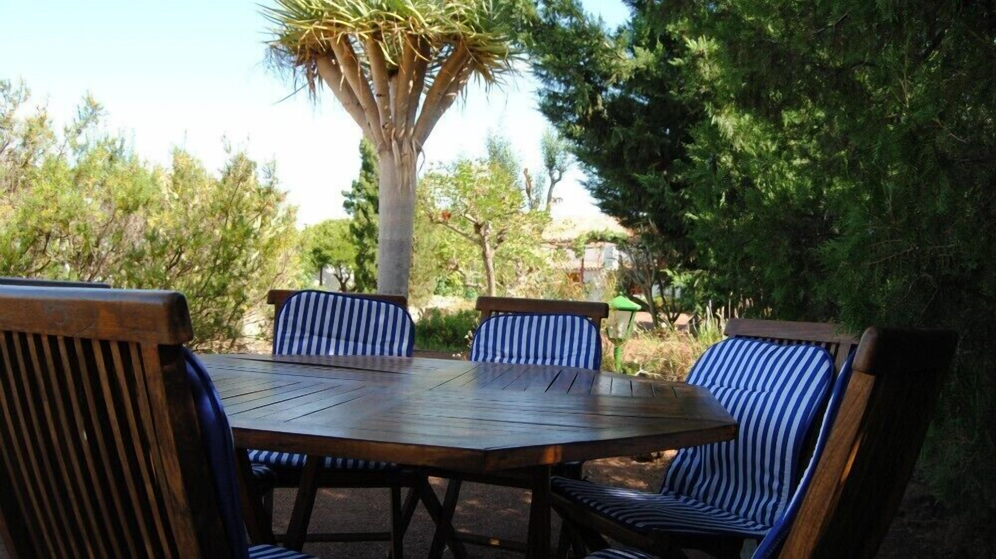 Un espace extérieur aménagé avec une table ronde en bois et des chaises à rayures, entouré d’arbres et de verdure.