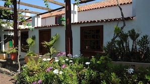 Un bâtiment blanc avec un toit recouvert de tuiles, des portes en bois et des fenêtres avec des volets. On peut voir des plantes en pot et des fleurs au premier plan.