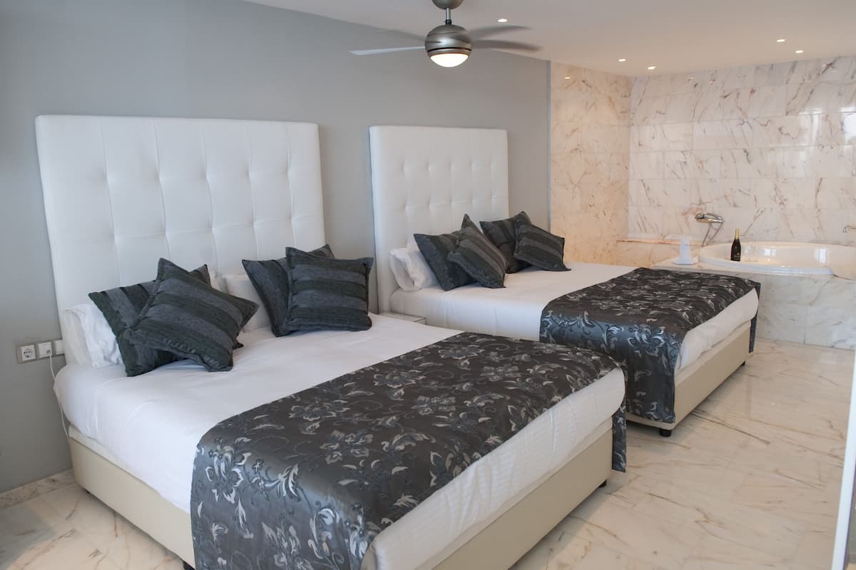 A modern bedroom with two beds, a large mirror, a ceiling fan, and a marble bathroom.