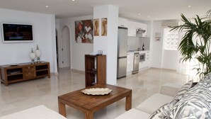 A modern living room with a wooden coffee table, a sofa, and a TV. There is a kitchen in the background with a refrigerator and a sink. - Club Puerto Atlantico (Mogan)