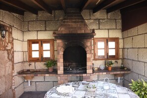 A stone fireplace with two windows, a table with a tiled tablecloth, and potted plants. - Casas Rurales Los Gavilanes (Arico)