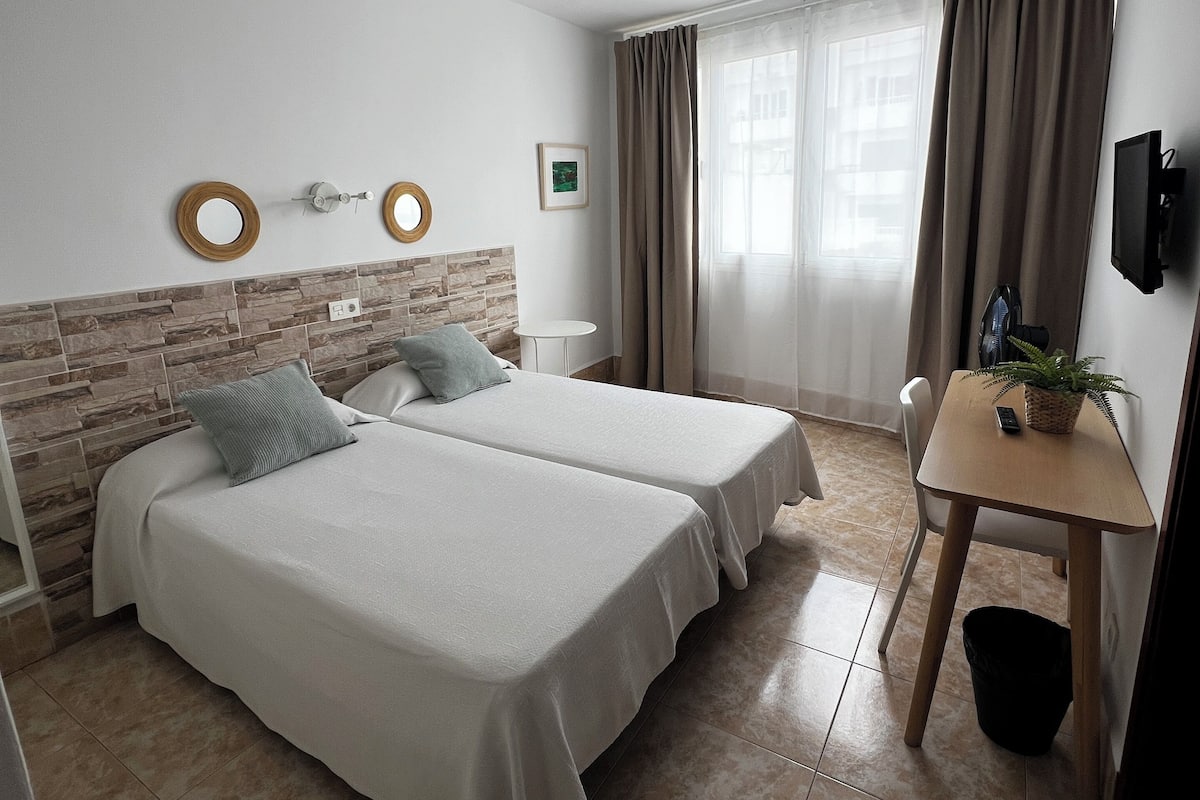Habitación de hotel con dos camas, un escritorio pequeño, televisión y ventana con cortinas.