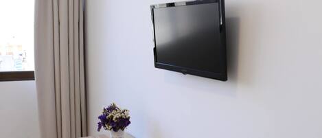 A room with a flat-screen TV mounted on the wall, a wooden table with a vase of flowers, a white chair, and a window with curtains.