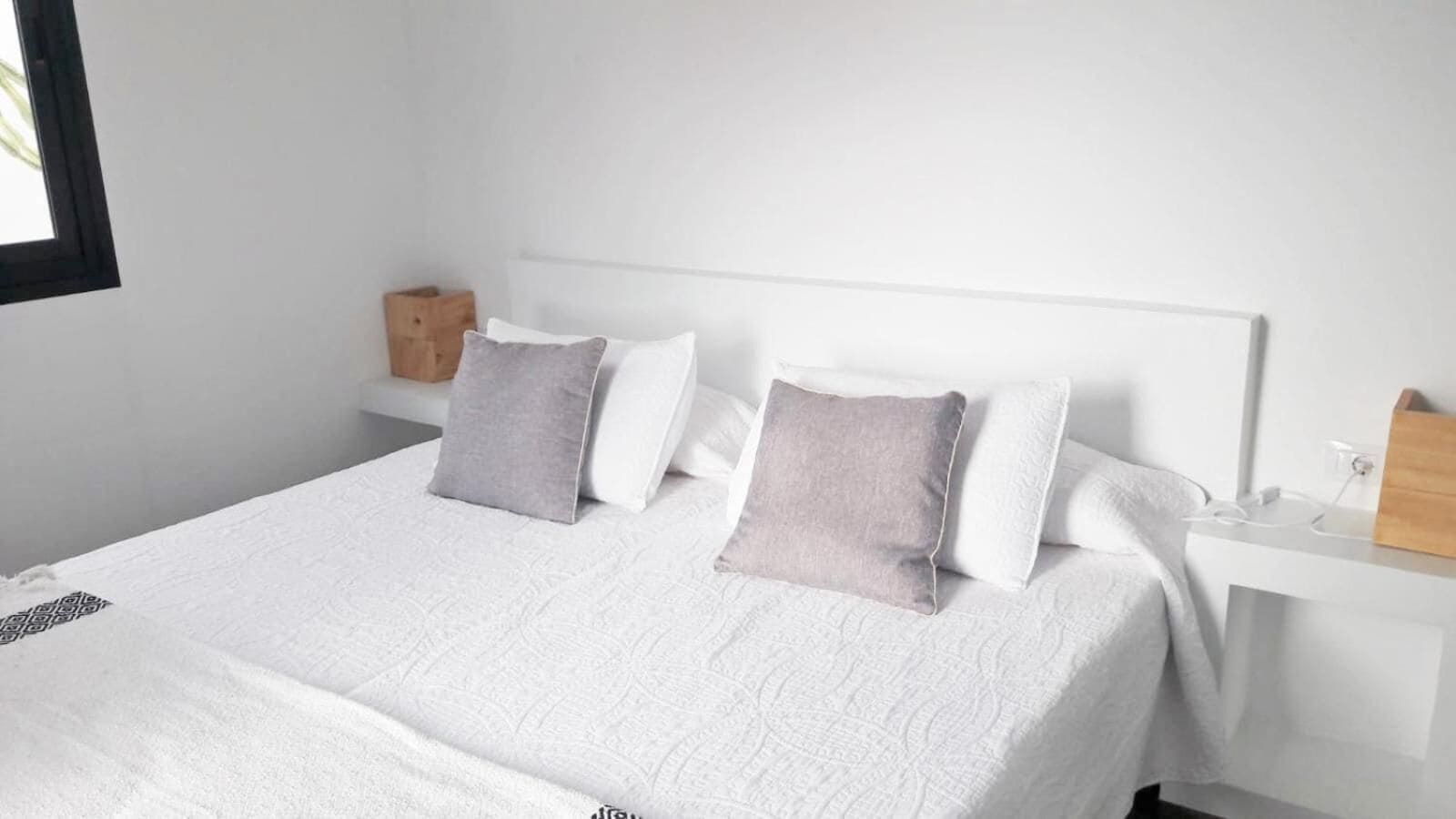 Una cama bien hecha con ropa blanca y dos almohadas, una mesita de noche con una lámpara y una pequeña mesita auxiliar de madera.