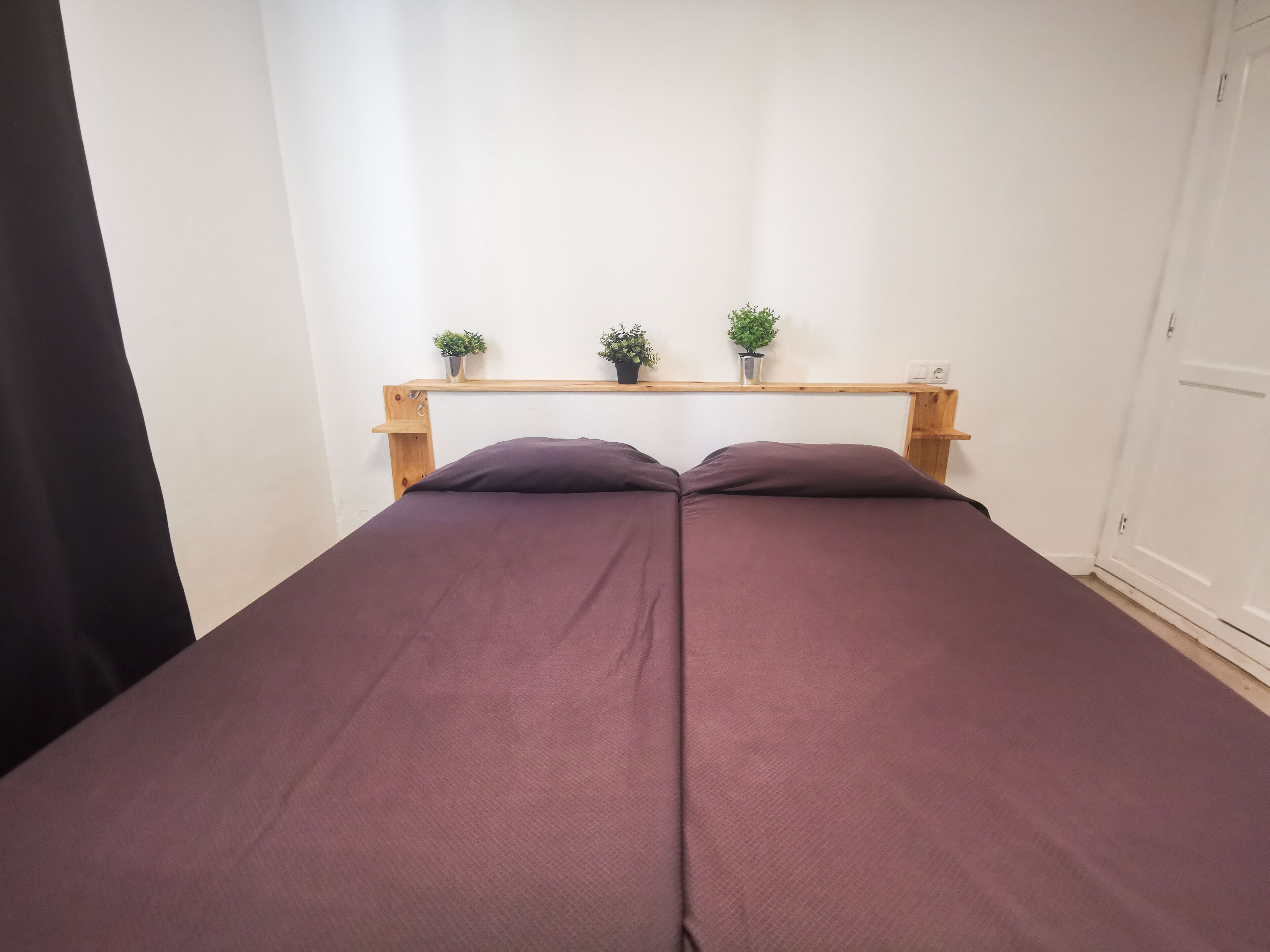 A bed with a wooden headboard, two potted plants on a shelf above, and a white door to the right.