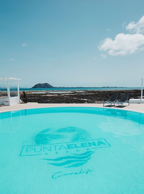 A clear blue pool with "PUNTADELMA" and "Costa" written on the surface, surrounded by lounge chairs and a white pergola, with a view of the ocean and distant land. - Punta Elena Beach (La Oliva)