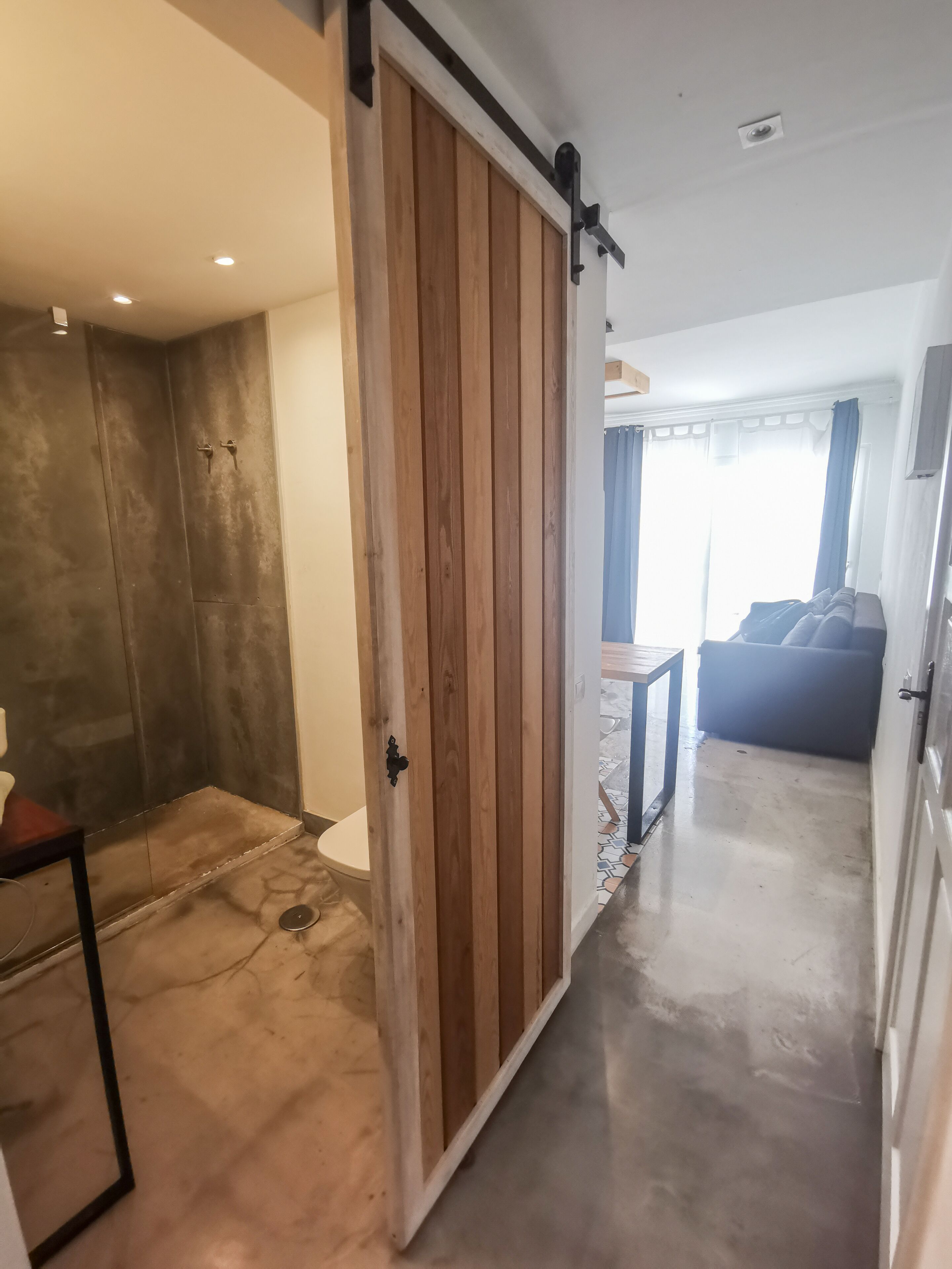 Un intérieur moderne avec une porte coulissante de style grange donnant sur une salle de bain, un coin repas avec une table et des chaises, et un salon avec un canapé et une vue sur l’extérieur.