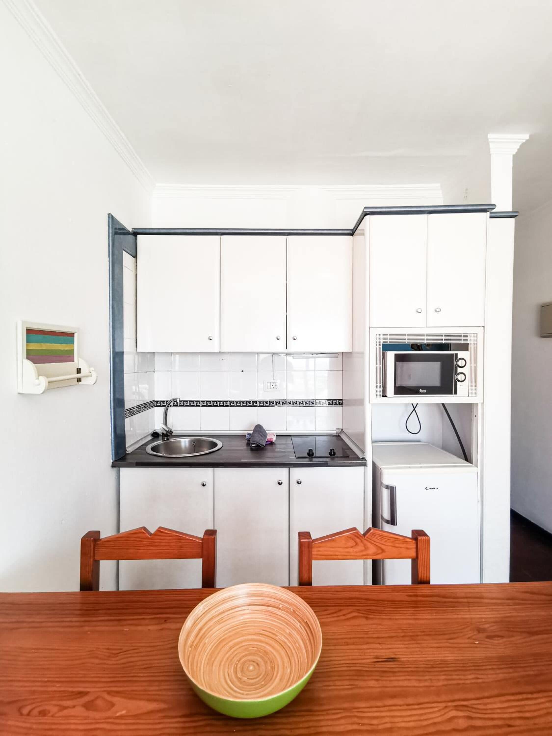 A compact kitchen with white cabinets, a microwave, and a sink. There is a wooden table with a green bowl in front.