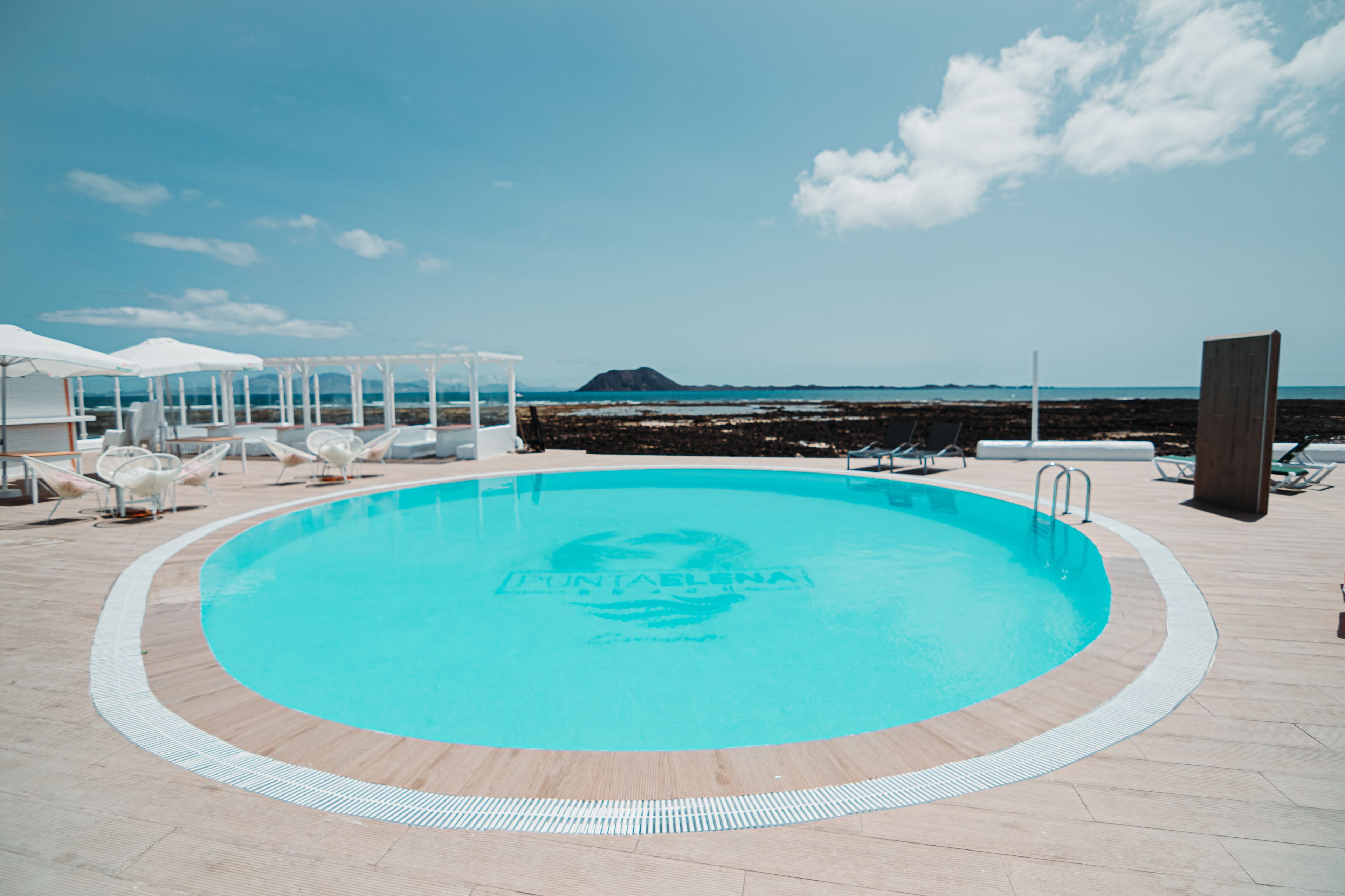 Un área de piscina con sillas de descanso y sombrillas, cielo despejado y vista al mar.