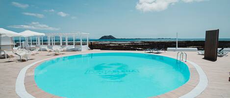 Un área de piscina con sillas de descanso y sombrillas, cielo despejado y vista al mar.