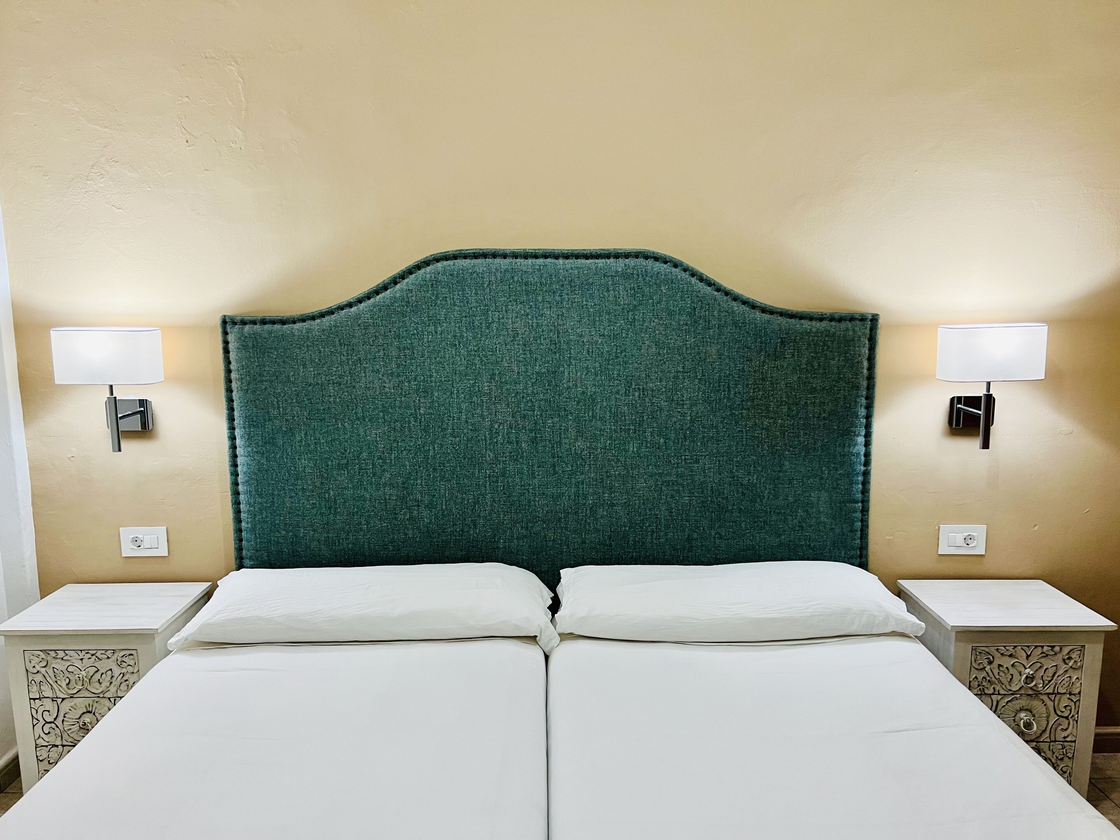 A double bed with a green upholstered headboard, two bedside tables with lamps, and a plain wall.