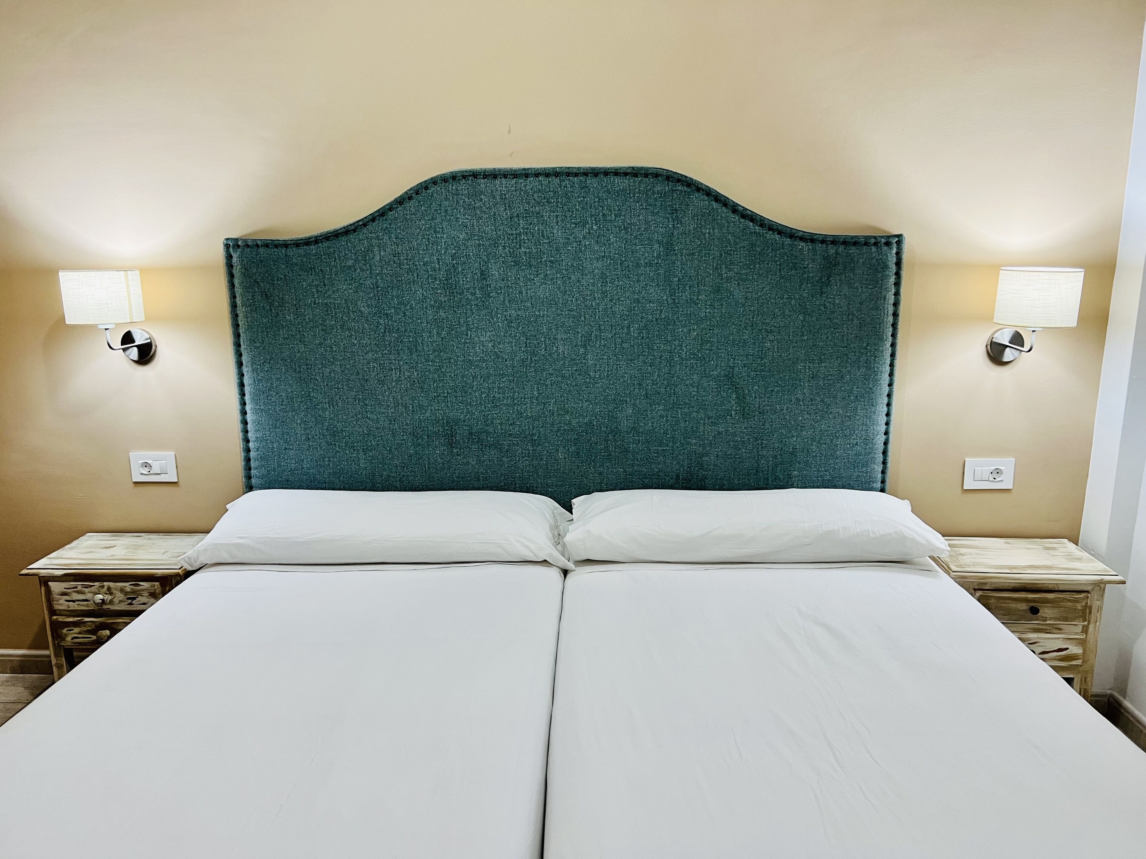 Una cama doble con sábanas blancas, cabecera color turquesa y dos mesitas de noche con lámparas.
