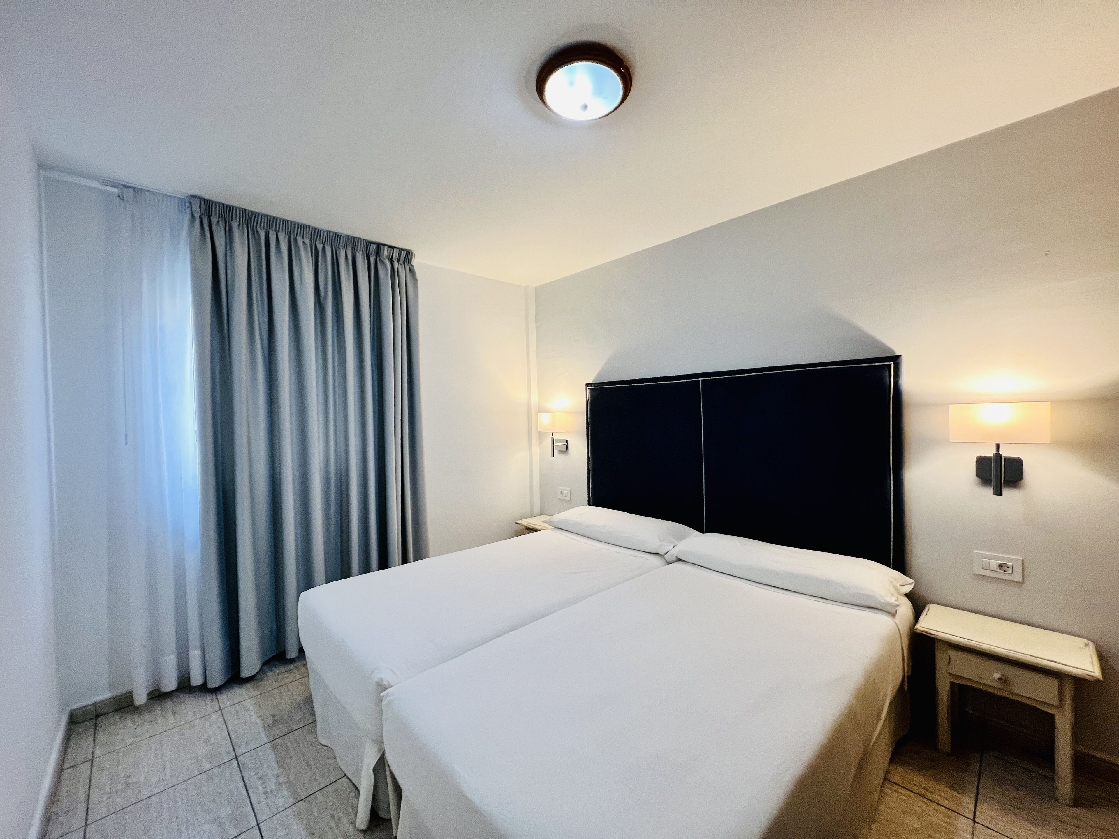 A hotel room with a large bed, a nightstand, a lamp, and a window with curtains.