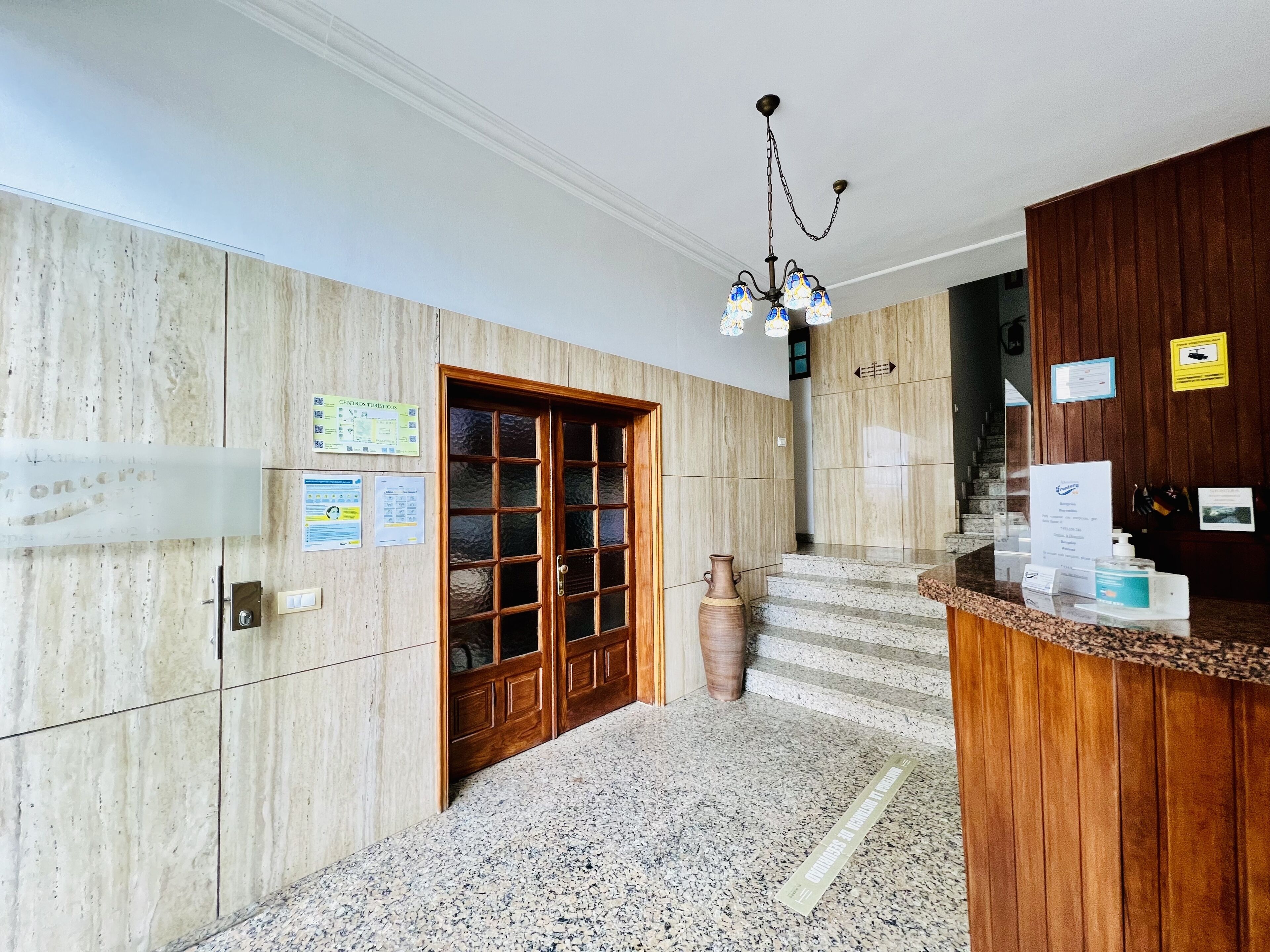 A hotel lobby with a wooden reception desk, a large wooden door, and a staircase leading to an upper floor.