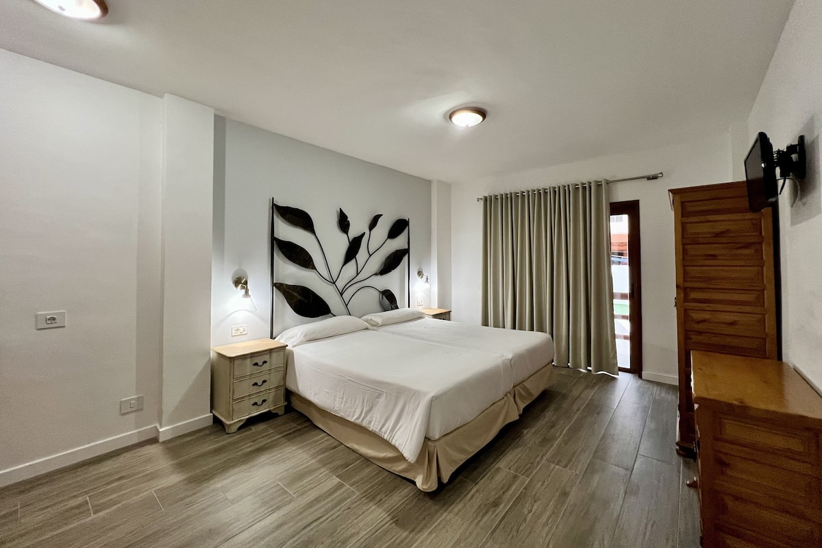 A bedroom with a large bed, a nightstand, a wall-mounted lamp, and a wooden wardrobe.