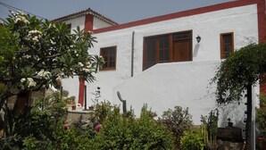 A white building with red trim and multiple windows, surrounded by lush greenery and flowering plants. - Casa Rural Anton Piche (Granadilla de Abona)