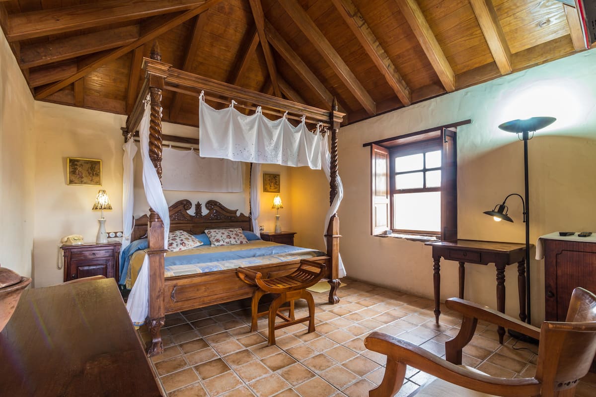 A traditional bedroom with a four-poster bed, wooden furniture, and a tiled floor.