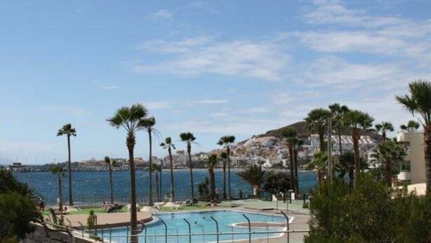 Un área de piscina con palmeras, vista al mar y edificios al fondo.
