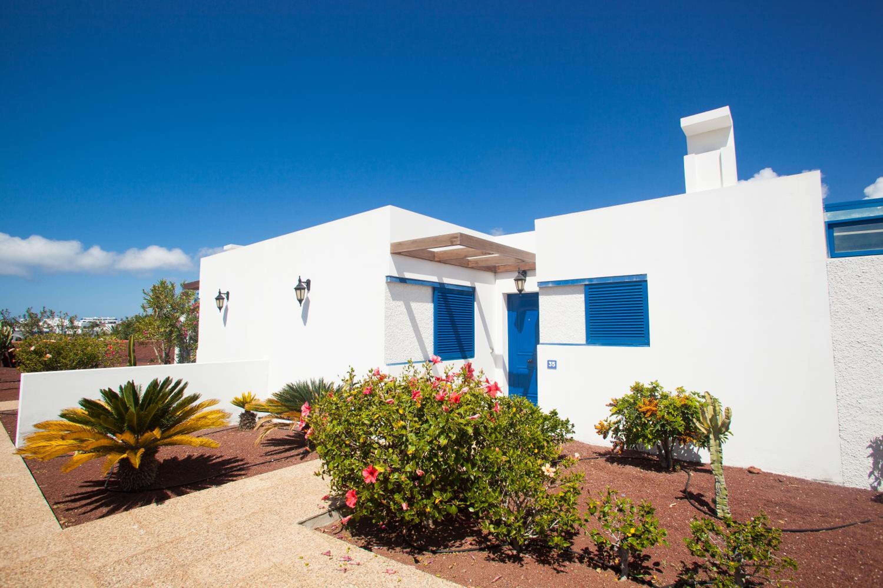 Un edificio moderno blanco con contraventanas azules, un pequeño jardín con diversas plantas y un cielo azul despejado.