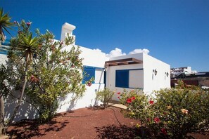 Um edifício moderno branco com persianas azuis, rodeado por vegetação exuberante e plantas floridas.