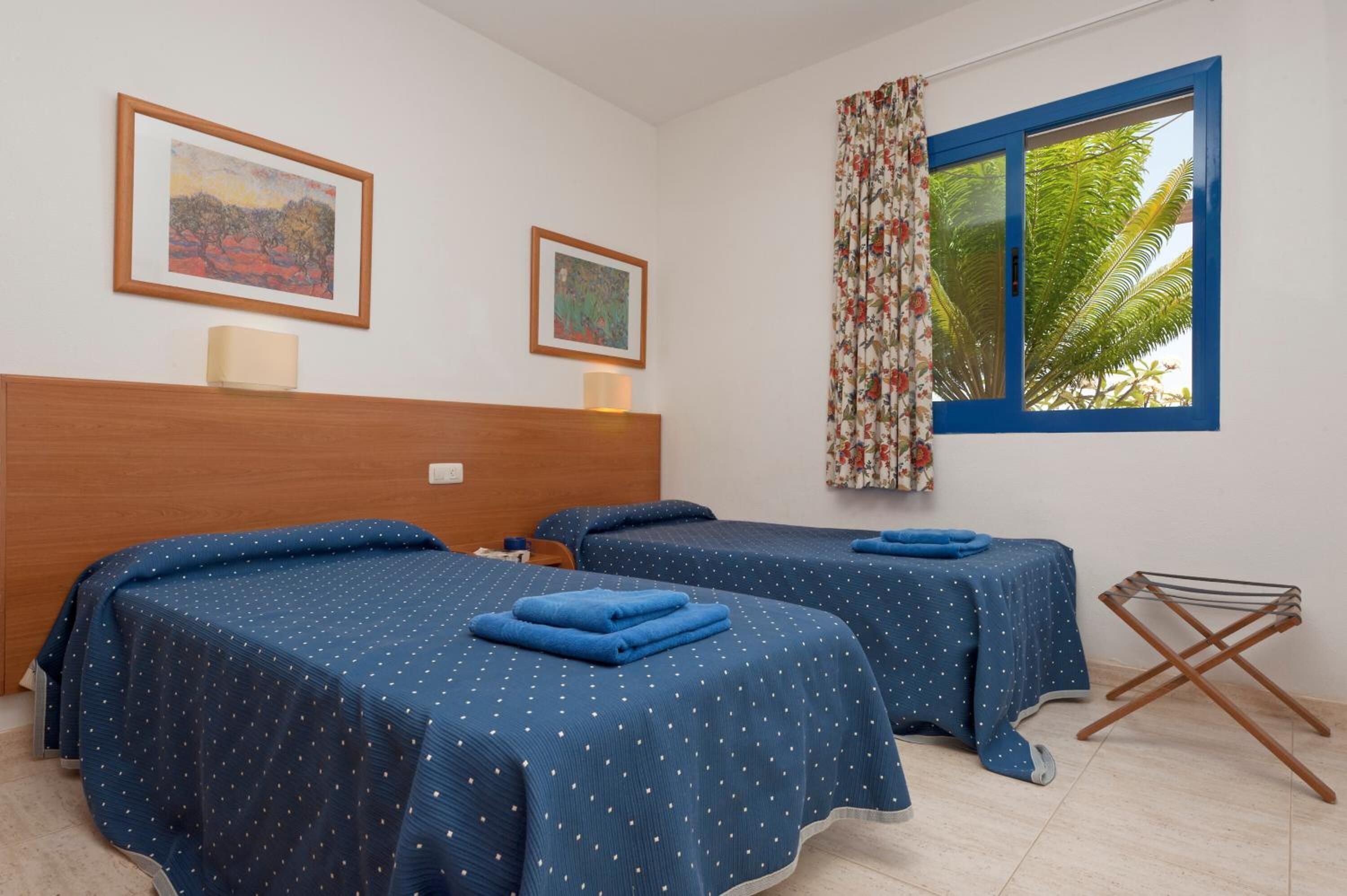 A hotel room with two beds, a wooden headboard, a window with curtains, and two framed artworks on the wall.