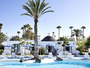 A pool area with lounge chairs and umbrellas, surrounded by palm trees and a building with a dome roof. - The Mur Bungalows Parque Romántico (San Bartolome de Tirajana)