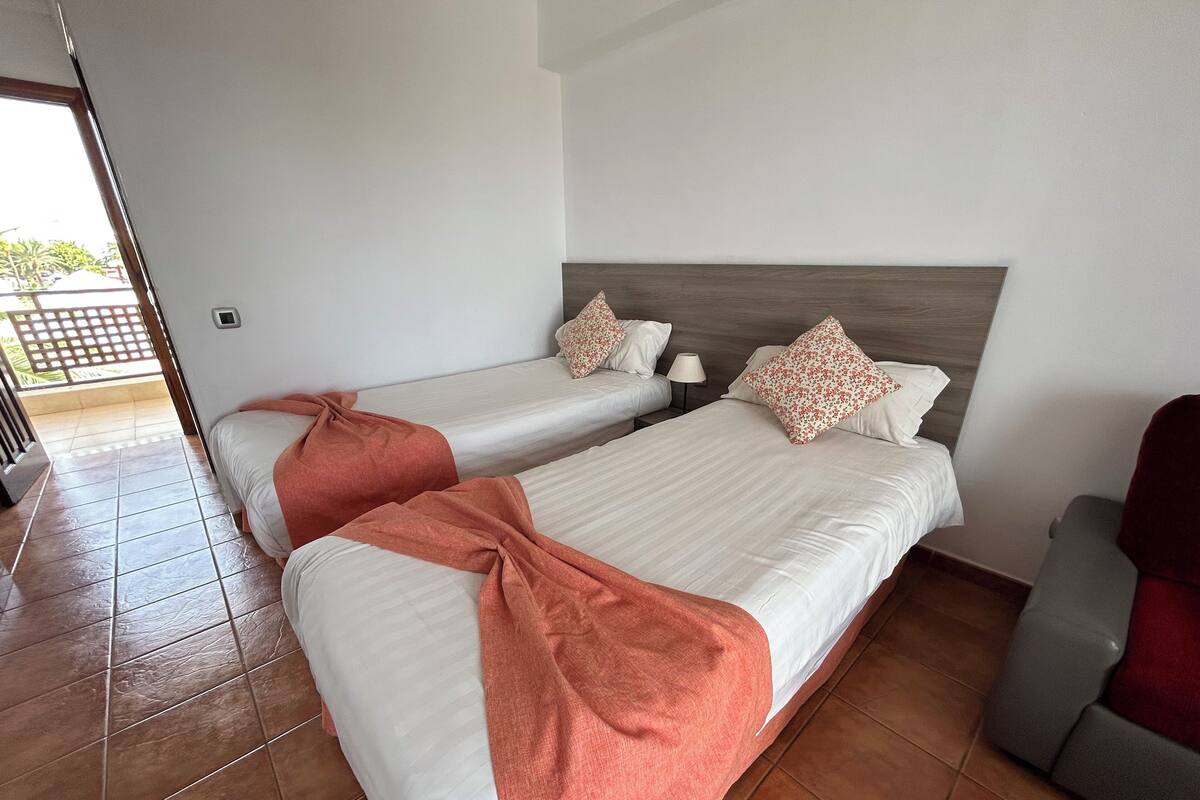 Una habitación de hotel con dos camas individuales, un cabecero de madera, una silla roja y suelo de baldosas.