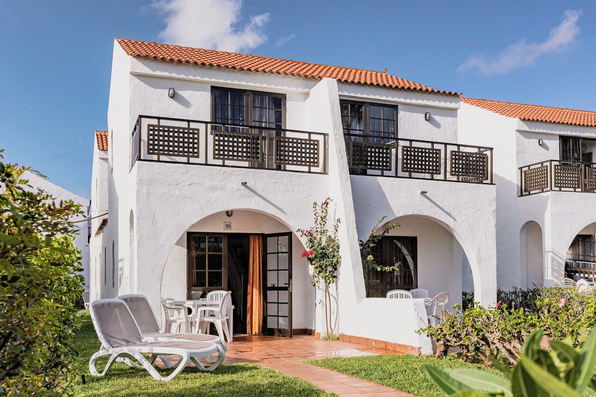 Un edificio blanco con techo de tejas, varios balcones y un patio empedrado con mobiliario exterior.