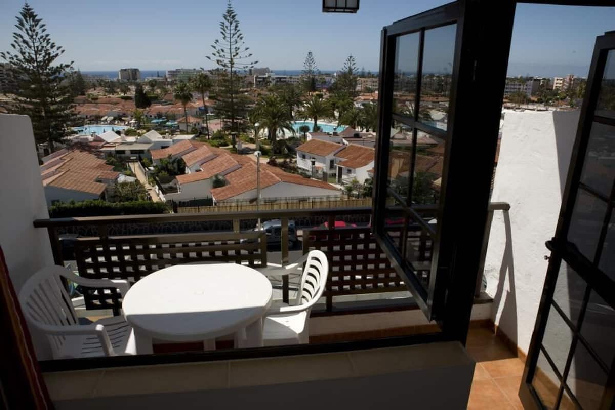 Un balcón con mesa y sillas, con vistas a una zona residencial con casas y cielo despejado.