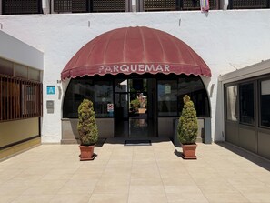 Un edificio con un toldo rojo que dice 'PARQUEMAR' y dos macetas con plantas en la entrada.