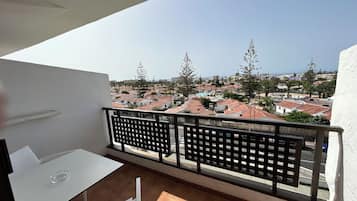 Un balcón con una mesa y sillas blancas, vista a una zona residencial con casas de techo rojo y árboles, y un cielo despejado.