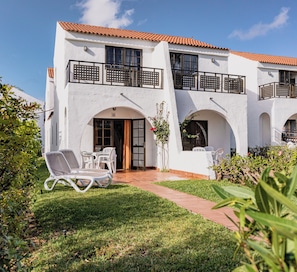 Un edificio blanco con techo de tejas, varios balcones y un patio empedrado con mobiliario exterior.