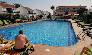 Una piscina con sillones de playa y sombrillas, rodeada de edificios con tejados rojos.