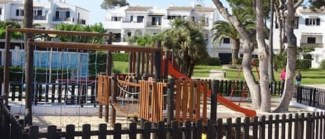 A playground with a wooden climbing structure and a slide, surrounded by a fence and palm trees.