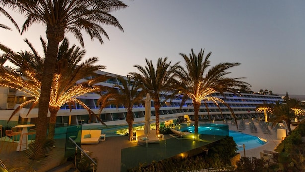 A hotel with a swimming pool, palm trees, and a modern building with illuminated windows. - Santa Mónica Suites Hotel (San Bartolome de Tirajana)
