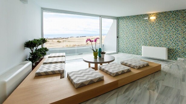 A modern living room with a large window, a round table with cushions, and a patterned wall. - Santa Mónica Suites Hotel (San Bartolome de Tirajana)