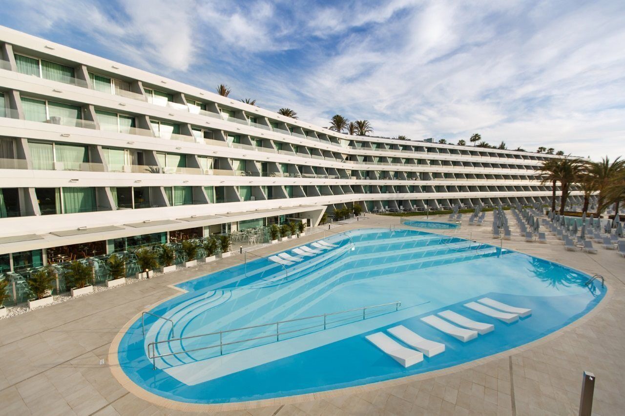 A modern hotel with a large pool area, multiple balconies, and a clear sky.