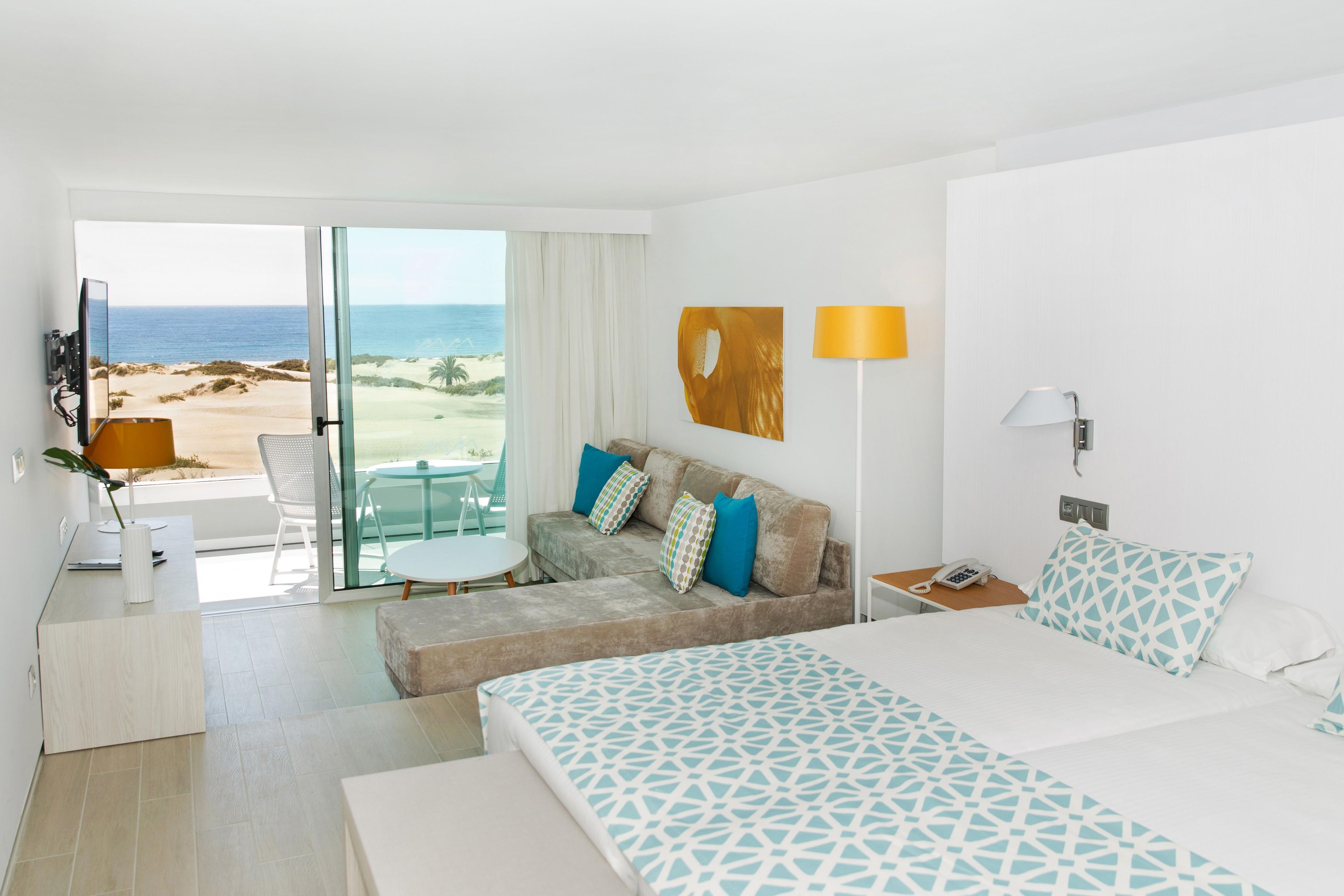 A hotel room with a bed, a sofa, a desk with a lamp, a TV, and a view of the beach.