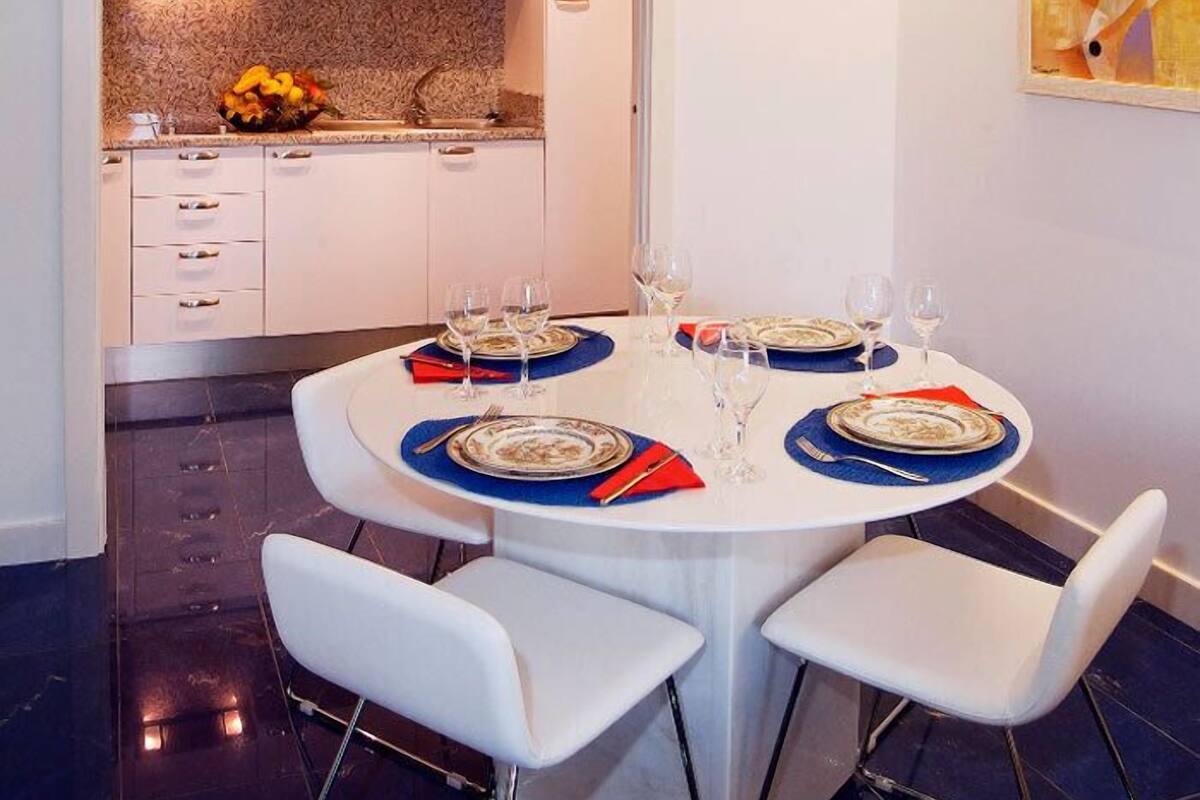 Uma sala de jantar com uma mesa redonda posta para quatro pessoas, cadeiras brancas e uma cozinha ao fundo.