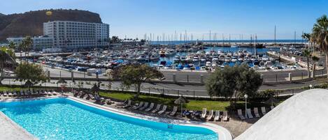 Een jachthaven met veel boten, een zwembad met ligbedden en een hotel op de achtergrond.
