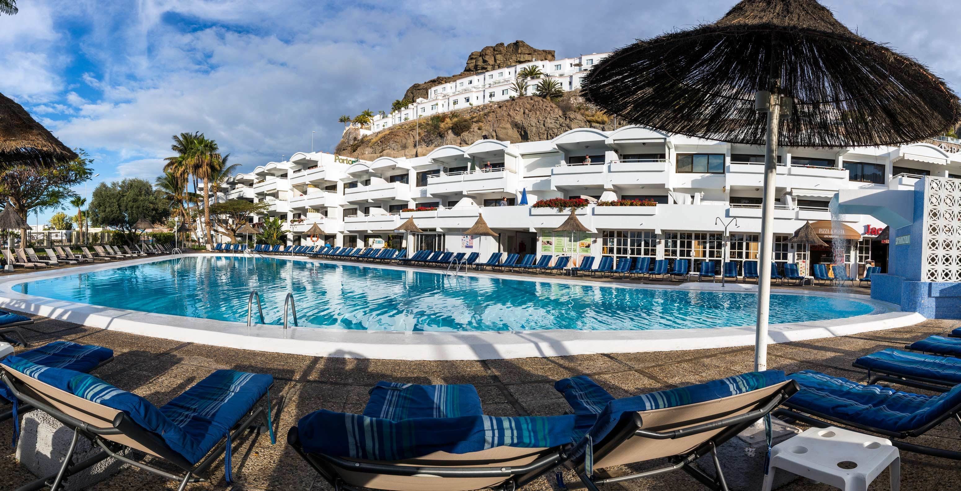 A hotel with a large pool surrounded by lounge chairs and umbrellas, white buildings with balconies, and a mountainous backdrop.