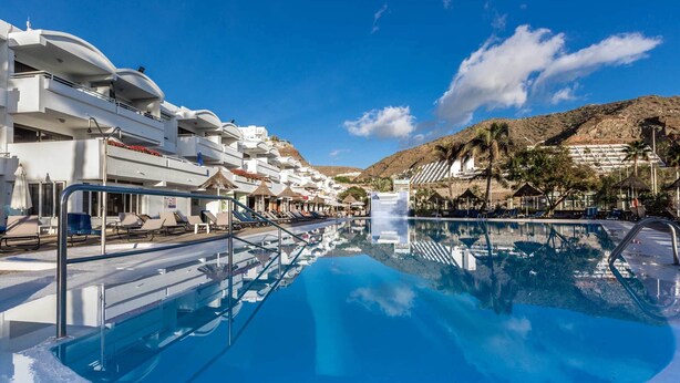 Une piscine entourée de chaises longues et de parasols, elle-même bordée de bâtiments blancs dotés de balcons, le tout sous un ciel dégagé.