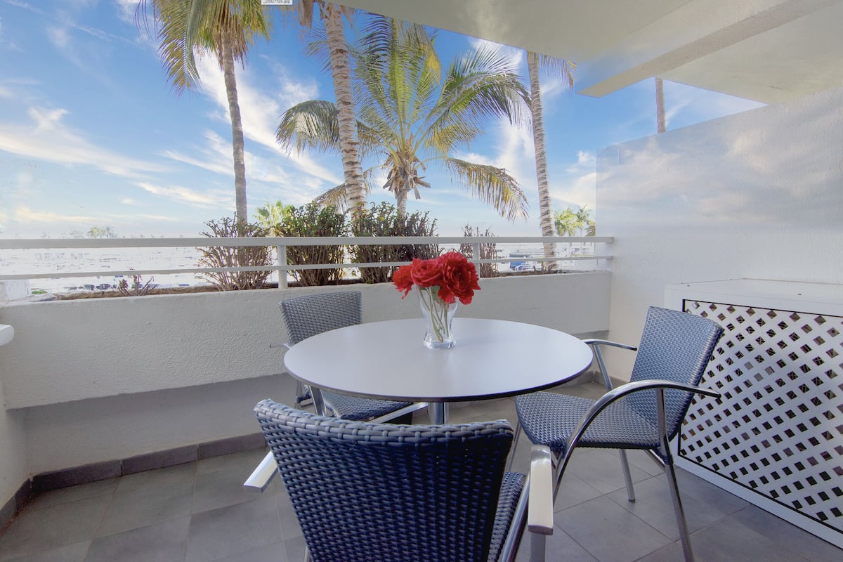 A round table with chairs and a vase of red flowers on a balcony with a view of palm trees and the sea.