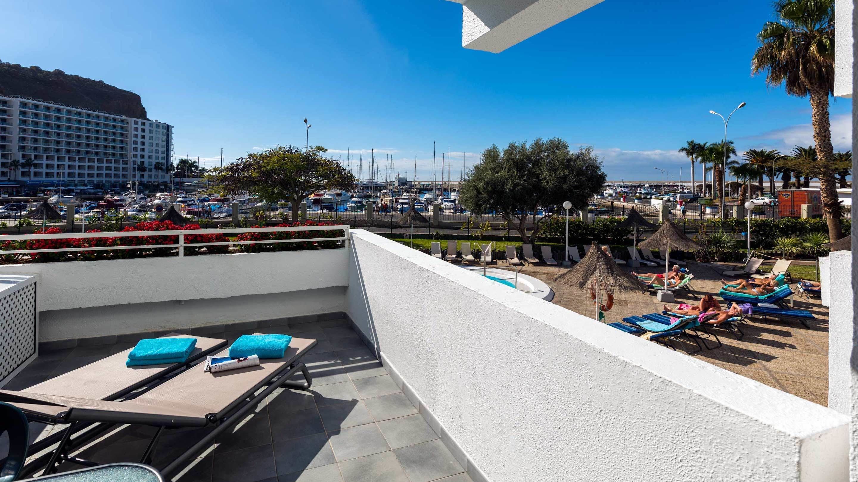 A rooftop terrace with lounge chairs and a pool, overlooking a marina with boats and a large hotel building.