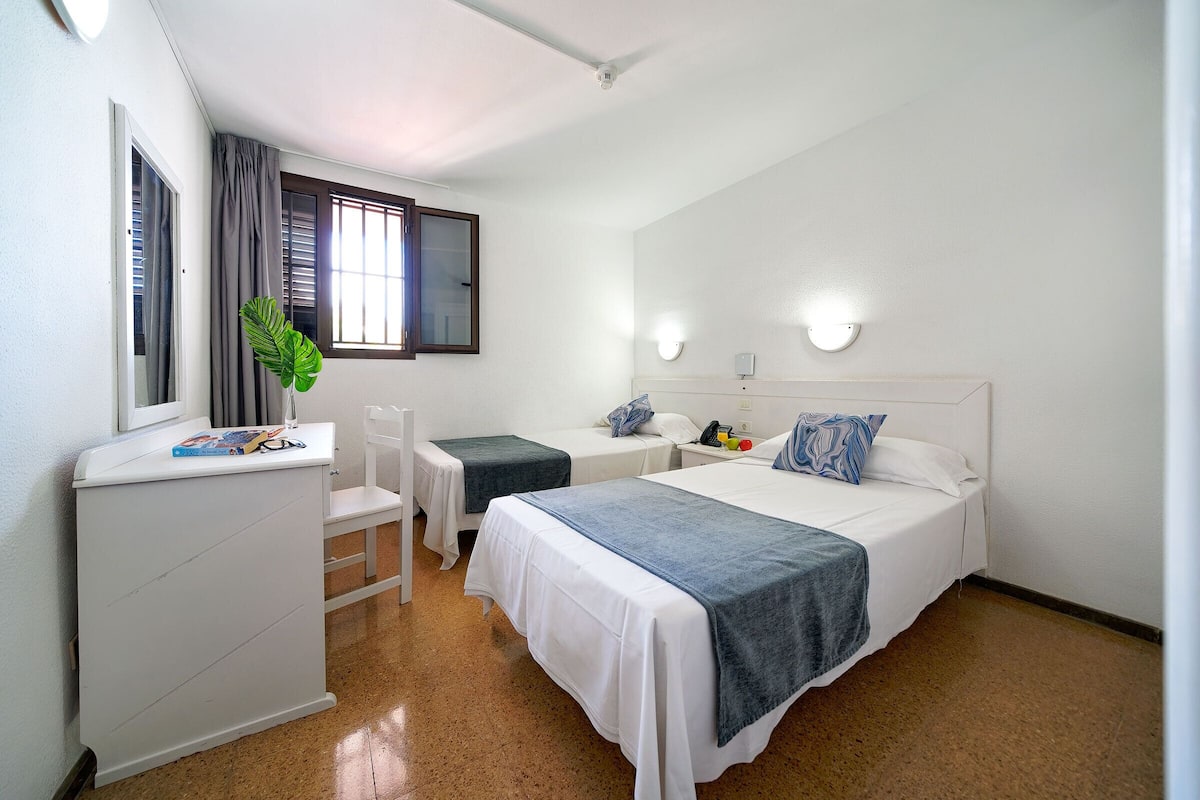 A hotel room with a bed, bedside tables, a desk, a chair, a mirror, a window with curtains, and a plant.