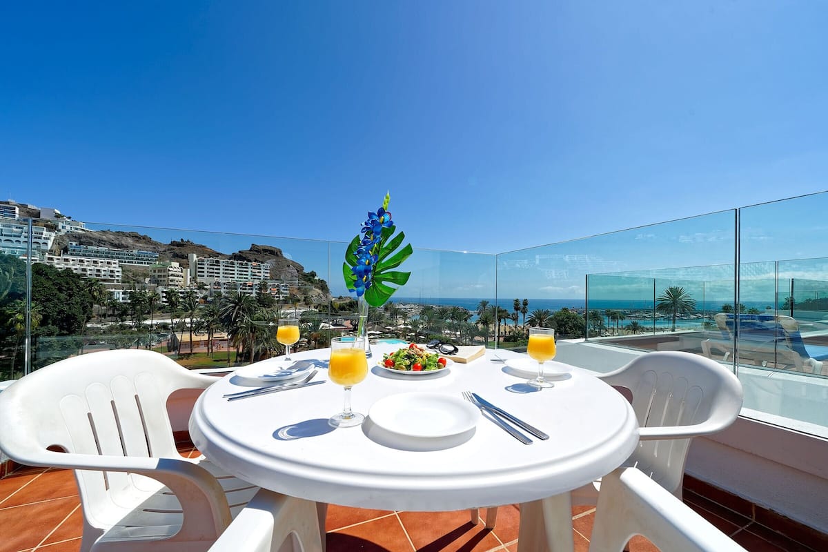 A table with a white tablecloth, two white plastic chairs, a plate of food, two glasses of orange juice, and a decorative blue flower on the table.